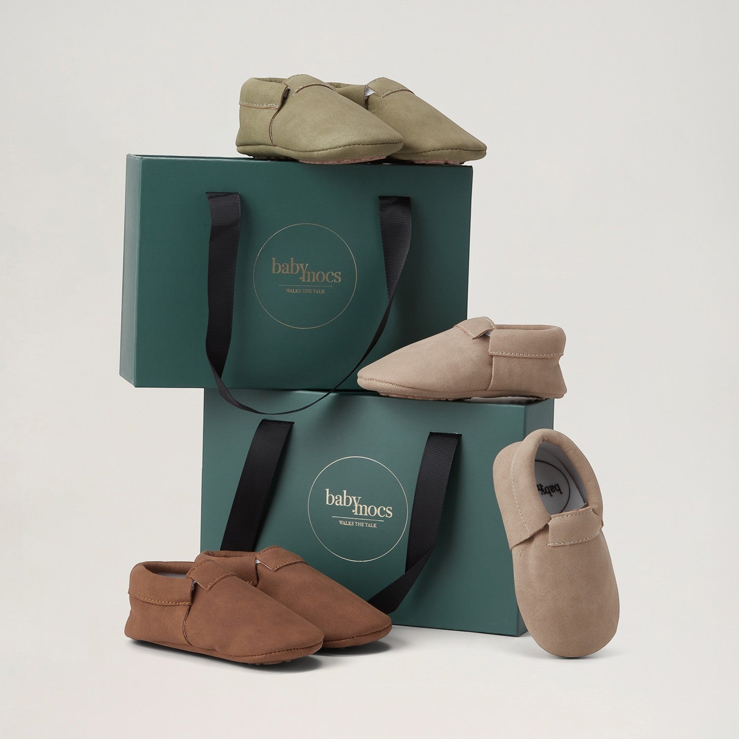 BabyMocs casual mocs för barn i olika nyanser av beige, brunt och grönt placerade på gröna presentförpackningar.